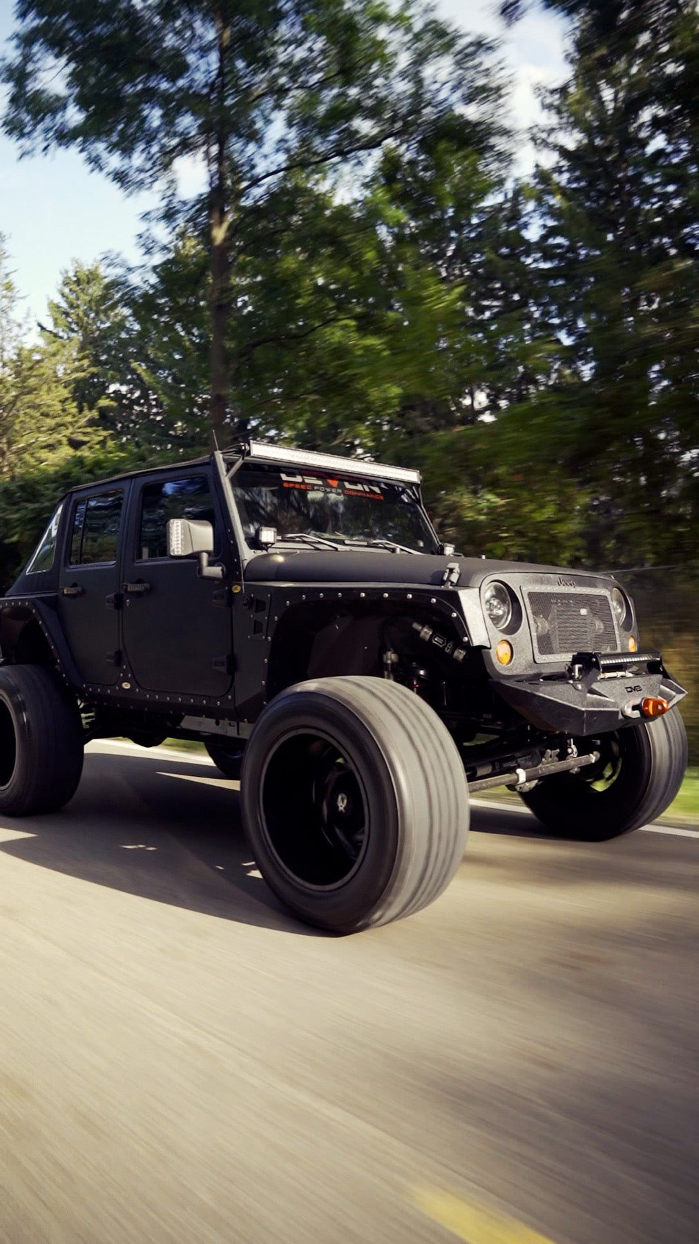 Devon Built custom blacked-out lifted Jeep Wrangler driving down a tree-lined road